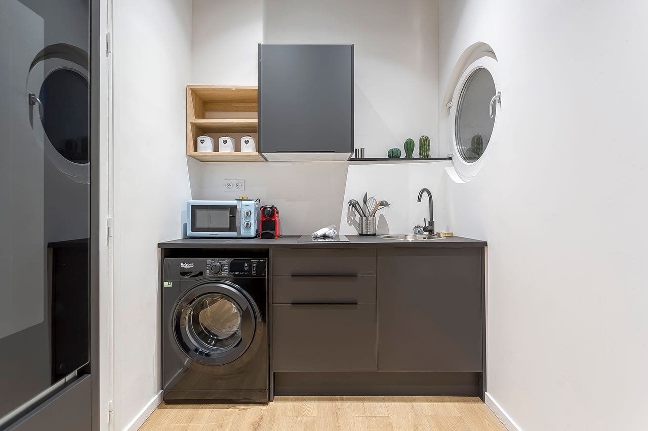 Kitchenette grise avec lave-linge sous plan, étagère en bois clair et fenêtre ronde de type hublot intégrée au mur, dans un studio rénové à Annemasse