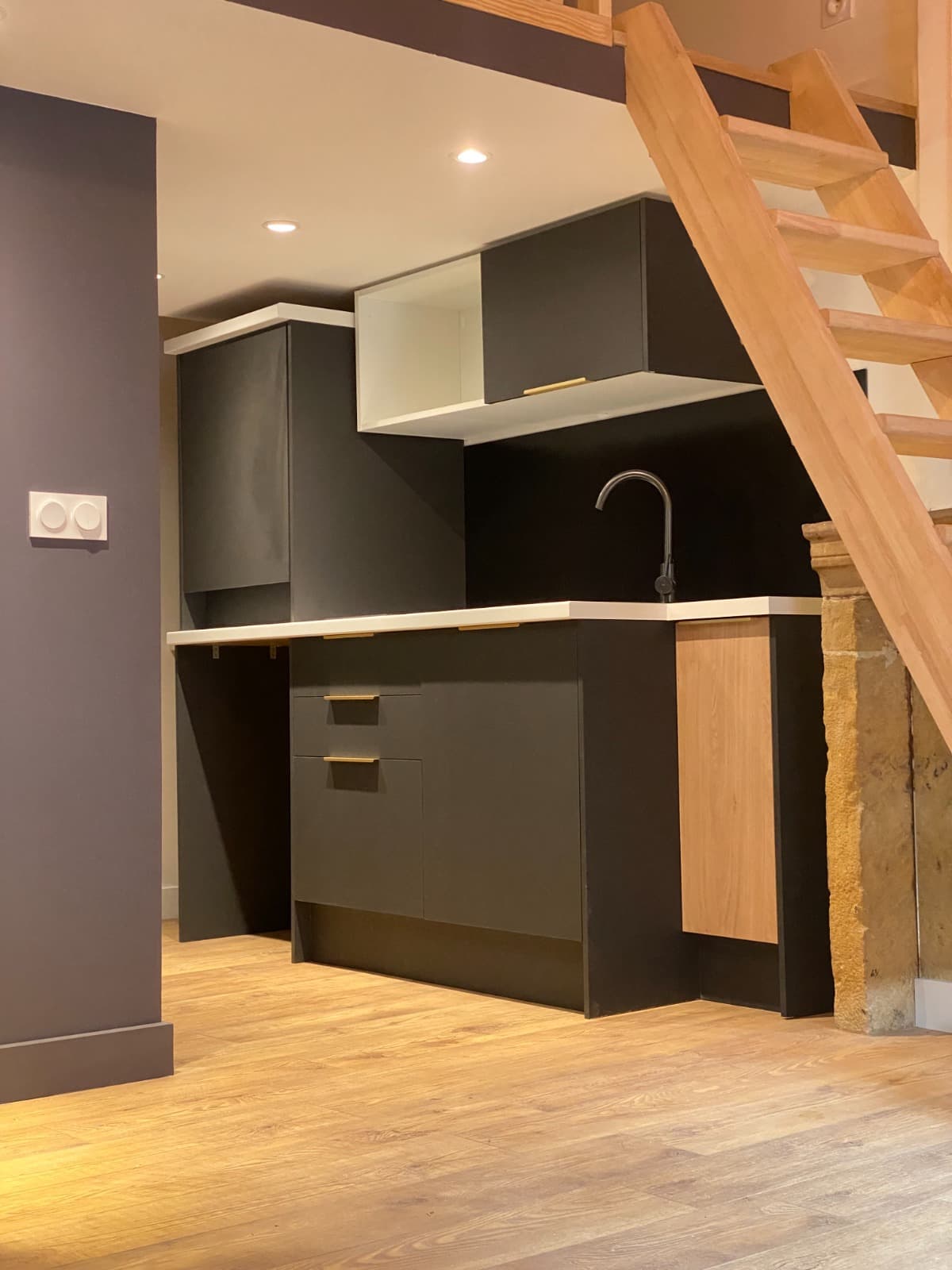 Kitchenette noire mate à poignées laiton et plan blanc installée sous la mezzanine, près d’un jambage de cheminée en pierre, dans un studio d’Annecy