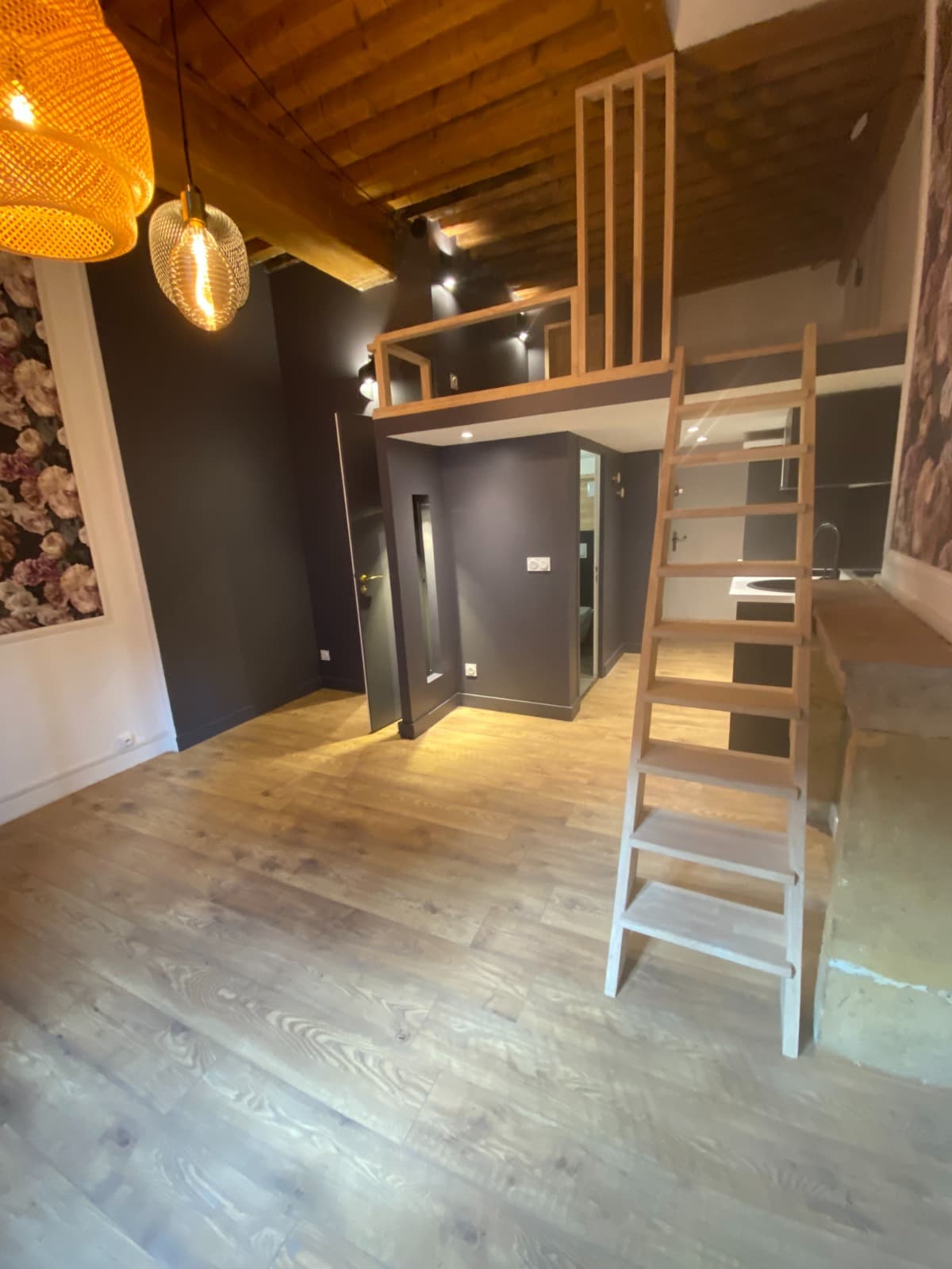 Studio rénové à Annecy : mezzanine bois à garde-corps à barreaux, plafond à solives apparentes et volume sous mezzanine en cloisons peintes anthracite